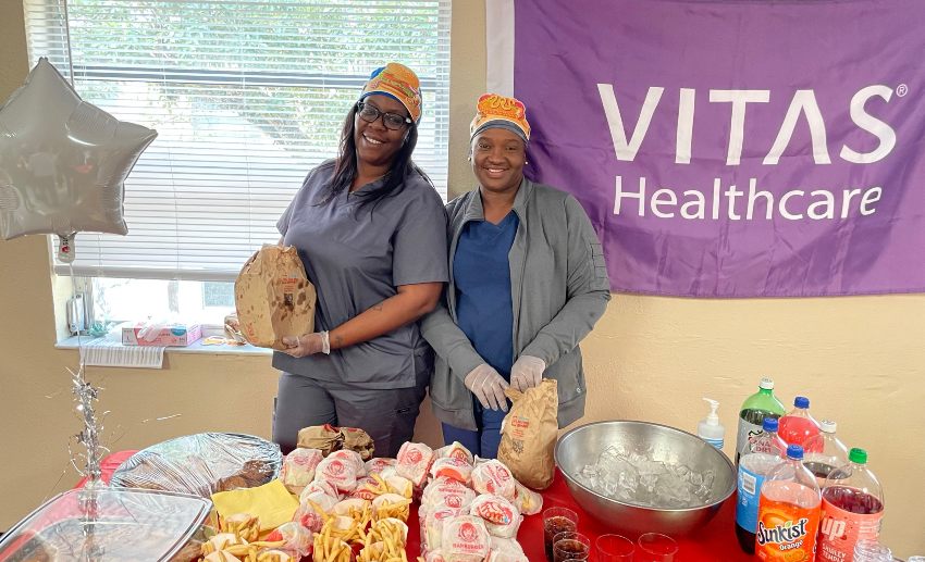 Two people stand behind a table of Wendy's hamburgers, French fries and 2-liter soda bottles with a purple VITAS Healthcare banner behind them