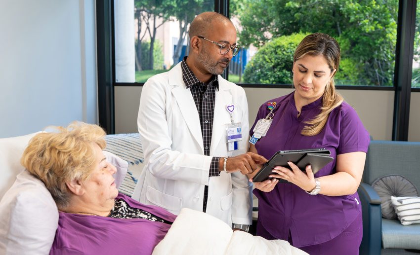 Two VITAS team members consult an iPad at an inpatient hospice unit as they talk with a patient in a hospital bed